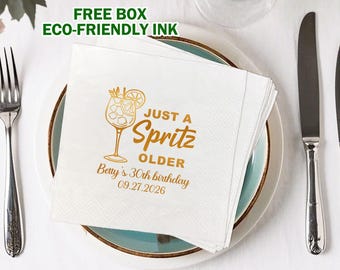 May include: A stack of white paper napkins with a gold-colored cocktail illustration and the text "JUST A Spritz OLDER Betty's 30th birthday 09.27.2026". The napkins are on a plate with a blue rim, next to silverware.