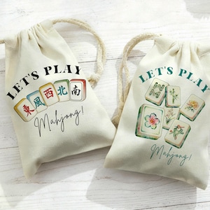May include: Two off-white drawstring bags with the text "LET'S PLAY Mahjong!" printed on them. One bag has colorful Mahjong tiles, and the other has floral Mahjong tiles. The bags have beige rope drawstrings.
