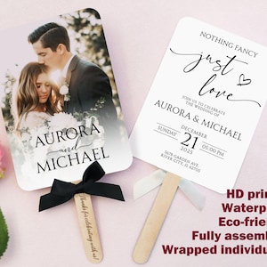 Puede incluir: Dos abanicos de boda blancos con mangos de madera. Un abanico presenta una foto de una pareja con los nombres "Aurora y Michael". El otro abanico tiene el texto "Nothing Fancy, just love" con detalles de la boda.