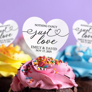 May include: White heart-shaped cupcake toppers with the text "Nothing Fancy Just Love" and the names Emily & David and the date November 17, 2025. The toppers are on top of cupcakes with pink, yellow, and blue frosting.