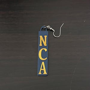 May include: A single navy blue earring with the letters NCA in gold. The earring is rectangular with rounded corners and a silver fish hook. The letters are in a bold, sans-serif font.