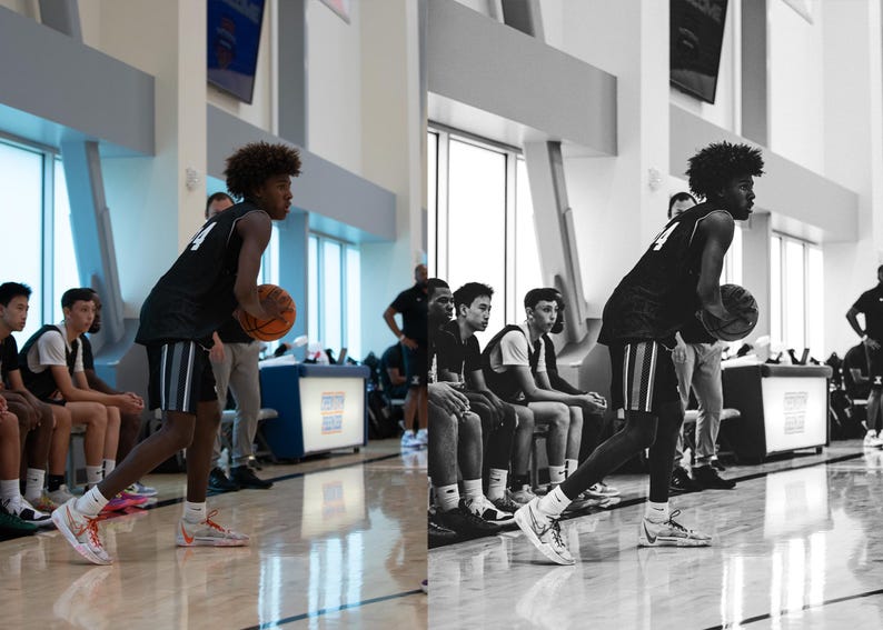 May include: A young man wearing a black and white basketball jersey dribbles a basketball while standing in a gym. Other young men are sitting on a bench in the background.