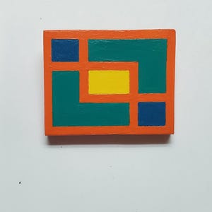 May include: A small, rectangular wooden block painted with an abstract geometric design. The design features a teal green background with a yellow rectangle in the center, surrounded by orange borders and blue accents.