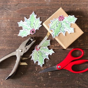 May include: A collection of holiday crafting supplies on a wooden surface. Includes a pair of red scissors, a silver hole punch, a small brown gift box, and three holly leaf cutouts with red berries.