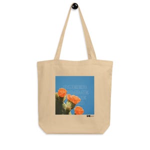 Könnte beinhalten: Beige Tragetasche mit einem quadratischen Grafikdesign, das einen Kaktus mit orangefarbenen Blüten vor blauem Hintergrund zeigt. Der Text "QUEER FEMME WILD" ist auf die Grafik gedruckt. Die Tasche hat einen schwarzen Griff.