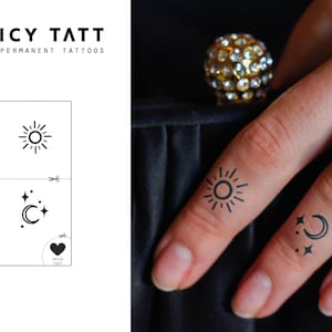 May include: A sheet of semi-permanent tattoos labelled JUICY TATT featuring black line art designs of a sun, a crescent moon, and stars. The sheet includes dotted cut lines and a heart icon. The tattoos are shown applied to two fingers.