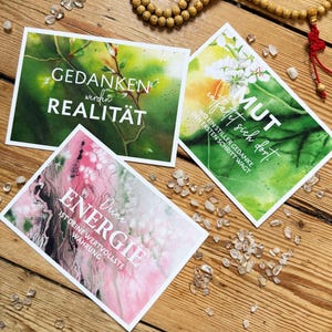 Drei Postkarten mit inspirierendem Text und Aquarellhintergründen werden auf einer Holzoberfläche zusammen mit einer Perlenkette und kleinen Kristallen präsentiert. Die Drucke zeigen Sätze wie "Gedanken werden Realität" , "Deine Energie ist deine wertvollste Währung" uns "Mut entfaltet sich dort, wo ein stiller Gedanke den ersten Schritt wagt" - Vision Board Set, Vision Board Ideen, Affirmationskarte