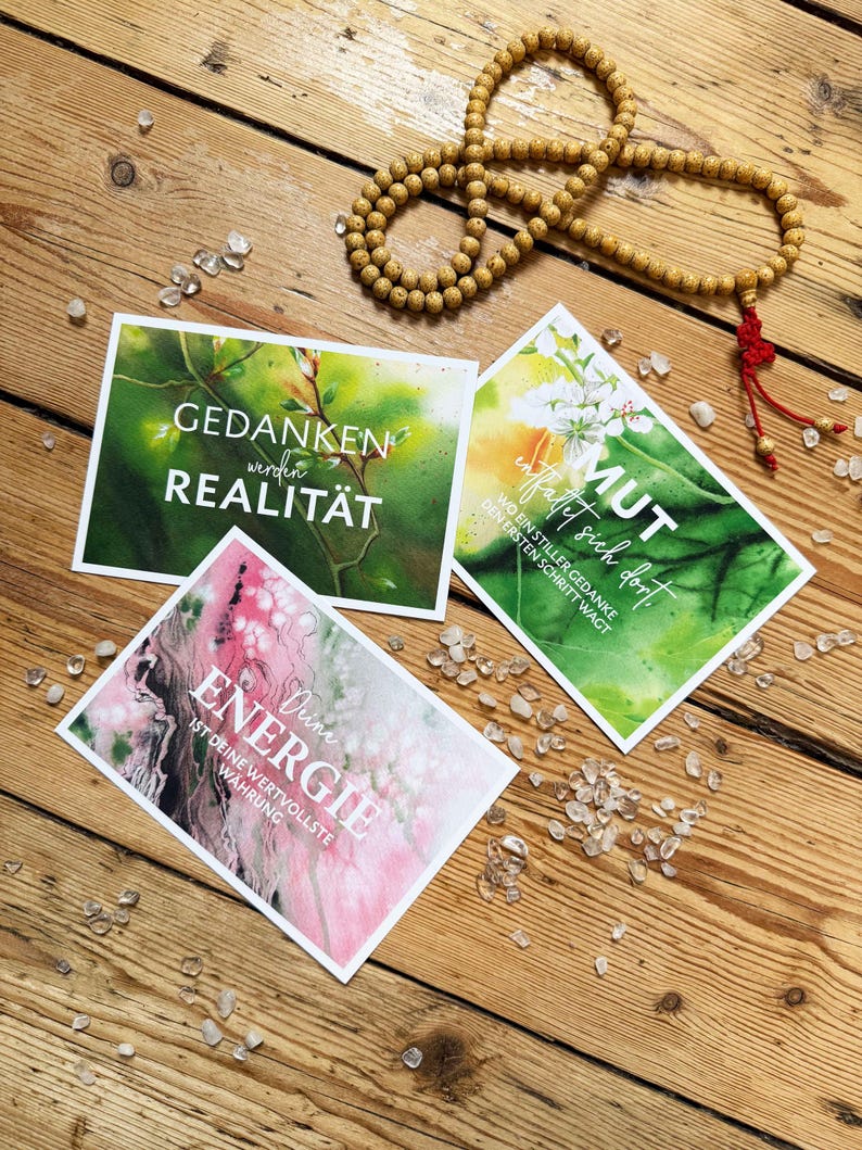 Drei Postkarten mit inspirierendem Text und Aquarellhintergründen werden auf einer Holzoberfläche zusammen mit einer Perlenkette und kleinen Kristallen präsentiert. Die Drucke zeigen Sätze wie "Gedanken werden Realität" , "Deine Energie ist deine wertvollste Währung" uns "Mut entfaltet sich dort, wo ein stiller Gedanke den ersten Schritt wagt" - Vision Board Set, Vision Board Ideen, Affirmationskarte