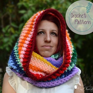 May include: A colorful rainbow crochet hooded scarf with a purple, blue, green, yellow, orange, and red color pattern. The scarf is worn by a person with red hair.