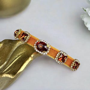 K&ouml;nnte beinhalten: Haarspange mit einem Senfgelben Samtband. Sie ist mit gro&szlig;en, ovalen und quadratischen, dunkelroten Edelsteinen verziert, die jeweils von kleinen, klaren Kristallen und goldfarbenem Metall umgeben sind.