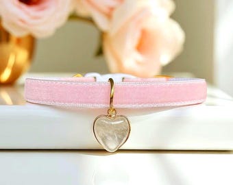 May include: A light pink velvet pet collar featuring a gold-toned metal ring and a heart-shaped pendant with a translucent, textured stone inlay. The collar includes a white plastic buckle closure and is displayed horizontally on a white surface.