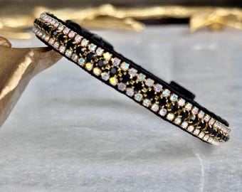 May include: Black pet collar with rows of iridescent and black rhinestones. The collar has a gold-colored chain design between the rows of rhinestones. The collar is on a light gray surface.