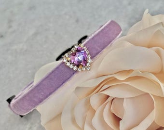 May include: A lavender velvet cat collar with a heart-shaped rhinestone charm. The charm features a purple crystal surrounded by smaller clear rhinestones.