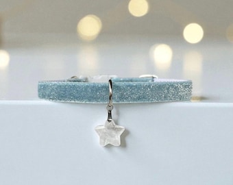 May include: A light blue velvet collar featuring a shimmering, glittery texture and a white plastic buckle. A silver-toned metal ring is attached to the collar, holding a small, translucent, five-pointed star charm that hangs vertically.