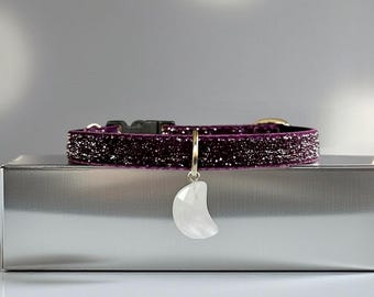 Könnte beinhalten: Ein dunkelviolettes Glitzerhalsband mit einer schwarzen Kunststoffschnalle und einem silberfarbenen Metallring. Ein weißer, durchscheinender Anhänger in Form eines Halbmonds hängt an dem Ring und bildet einen Blickfang am strukturierten Band.