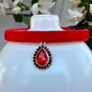 May include: A red velvet choker necklace with a gold pendant featuring a teardrop-shaped red gemstone surrounded by black rhinestones.