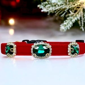 May include: A red velvet collar featuring three large, faceted emerald green gemstones set in gold-toned metal frames surrounded by small clear crystals. The collar includes a black plastic buckle closure and a gold-toned metal D-ring attachment.