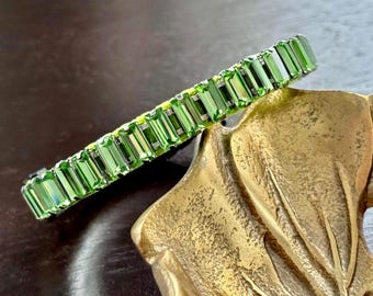 Puede incluir: Un brazalete verde con piedras preciosas de corte rectangular. El brazalete está hecho de múltiples piedras rectangulares verdes engastadas en un metal plateado. El brazalete descansa sobre un plato decorativo dorado.