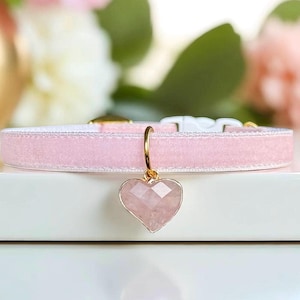 May include: A light pink velvet collar featuring a faceted heart-shaped pendant made of a translucent pink stone. The pendant is attached to the collar with a gold-toned metal ring. The collar includes a white plastic buckle closure mechanism.