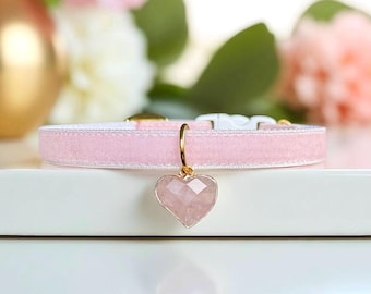 May include: A light pink velvet collar featuring a faceted heart-shaped pendant made of a translucent pink stone. The pendant is attached to the collar with a gold-toned metal ring. The collar includes a white plastic buckle closure mechanism.