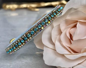 May include: A light blue headband adorned with rows of sparkling turquoise and iridescent crystals. The headband is set against a soft pink rose, highlighting its elegant design. A gold-colored metal loop is visible on the side.