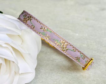 May include: A lavender ribbon with a gold and green floral pattern. The ribbon is curled and has a gold clasp.