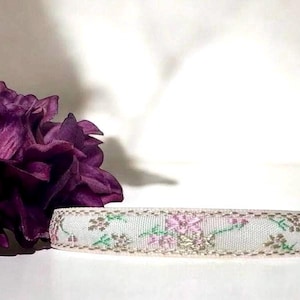 K&ouml;nnte beinhalten: Ein wei&szlig;es Band mit einem floralen Muster in Rosa, Gr&uuml;n und Gold. Das Band liegt auf einer wei&szlig;en Oberfl&auml;che.