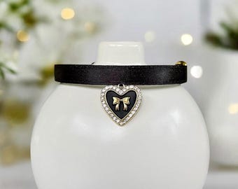 May include: Black satin pet collar with a heart-shaped charm. The charm features a black center with a gold bow and is surrounded by clear crystals. A gold-colored clasp secures the collar.