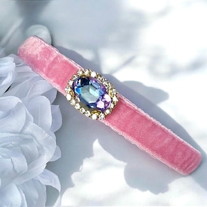 Puede incluir: Una diadema de terciopelo rosa con una gran piedra preciosa iridiscente de forma ovalada rodeada de diamantes de imitación transparentes en un engaste dorado.