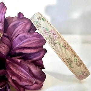 K&ouml;nnte beinhalten: Ein beigefarbenes Band mit einem floralen Muster in Rosa und Gr&uuml;n. Das Band ist gerollt und hat einen gen&auml;hten Rand.
