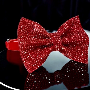 May include: A red bow tie made of sparkling rhinestones. The bow tie is on a black surface.
