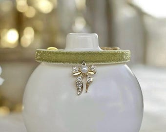 May include: A light green glittery fabric choker necklace featuring a central gold-toned bow charm. The bow is embellished with small clear crystals and a single white faux pearl at its center, suspended from the textured green band.