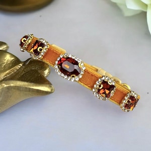 K&ouml;nnte beinhalten: Ein goldfarbenes Armband mit einem senffarbenen Samtband. Das Armband hat drei quadratische und einen ovalen, dunkelroten Edelstein, die jeweils von kleinen, klaren Kristallen umgeben sind. Das Armband ist vor einem wei&szlig;en Hintergrund platziert.