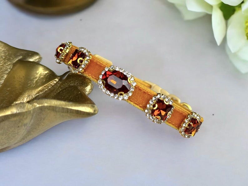 K&ouml;nnte beinhalten: Ein goldfarbenes Armband mit einem senffarbenen Samtband. Das Armband hat drei quadratische und einen ovalen, dunkelroten Edelstein, die jeweils von kleinen, klaren Kristallen umgeben sind. Das Armband ist vor einem wei&szlig;en Hintergrund platziert.