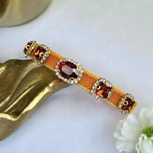 K&ouml;nnte beinhalten: Ein orangefarbenes Samt-Haarband, verziert mit gro&szlig;en, ovalen und quadratischen roten Edelsteinen. Die Edelsteine sind von kleinen, klaren Kristallen und goldfarbenen Metalldetails umgeben. Das Haarband wird auf einer wei&szlig;en Oberfl&auml;che pr&auml;sentiert.