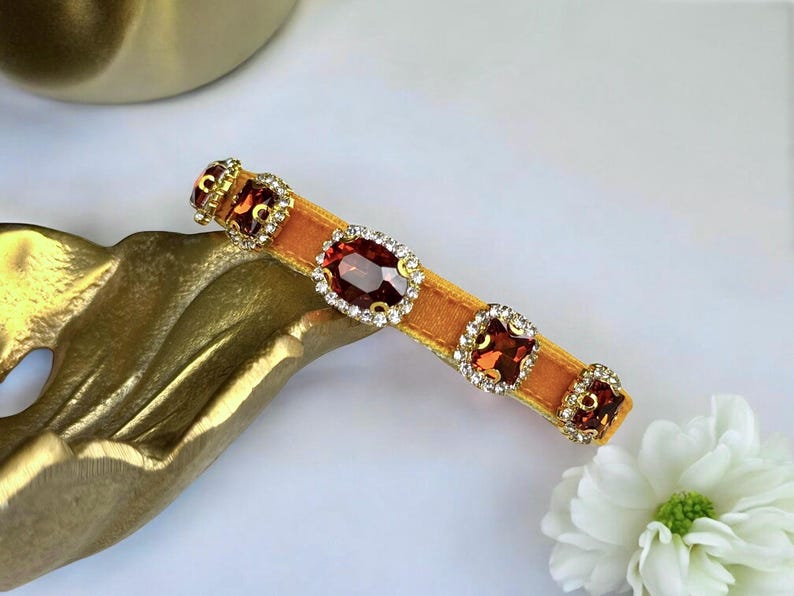 K&ouml;nnte beinhalten: Ein orangefarbenes Samt-Haarband, verziert mit gro&szlig;en, ovalen und quadratischen roten Edelsteinen. Die Edelsteine sind von kleinen, klaren Kristallen und goldfarbenen Metalldetails umgeben. Das Haarband wird auf einer wei&szlig;en Oberfl&auml;che pr&auml;sentiert.