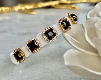 May include: A white headband adorned with five black and gold square embellishments. Each square is outlined with small, sparkling crystals. The headband is displayed on a white surface, with a blurred background.