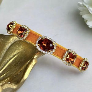K&ouml;nnte beinhalten: Orangefarbene Haarspange aus Samt, verziert mit gro&szlig;en, funkelnden roten Edelsteinen und kleineren klaren Kristallen. Die Spange hat eine goldfarbene Metalleinfassung f&uuml;r elegante Frisuren.