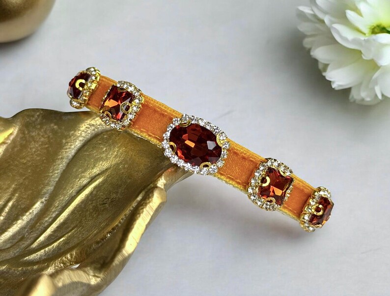 K&ouml;nnte beinhalten: Orangefarbene Haarspange aus Samt, verziert mit gro&szlig;en, funkelnden roten Edelsteinen und kleineren klaren Kristallen. Die Spange hat eine goldfarbene Metalleinfassung f&uuml;r elegante Frisuren.