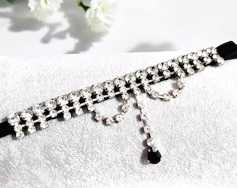 May include: A black velvet choker necklace adorned with two rows of sparkling clear rhinestones. A dangling strand of rhinestones ends with a black teardrop-shaped gem. The necklace is a glamorous accessory.