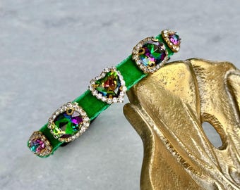 Puede incluir: Un collar gargantilla de terciopelo verde con detalles de joyas iridiscentes. Las joyas están engastadas en metal dorado y rodeadas de diamantes de imitación transparentes.