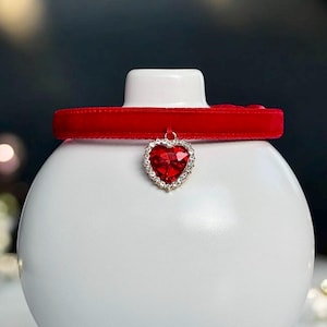 Puede incluir: Un collar de terciopelo rojo con un colgante en forma de corazón que presenta un strass rojo rodeado de strass transparentes.