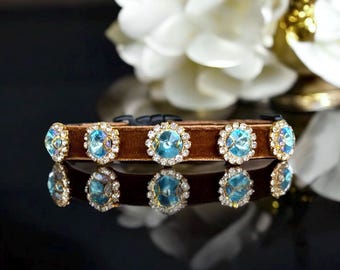 Puede incluir: Un collar de terciopelo marrón para mascotas adornado con grandes adornos de cristal azul claro y dorado. Los cristales están rodeados de cristales más pequeños y transparentes. El collar se refleja en una superficie negra brillante.