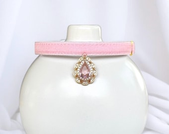 Puede incluir: Un collar rosa de terciopelo para mascotas con un colgante dorado. El colgante tiene forma de lágrima, incrustado con pequeños cristales transparentes y una gran piedra rosa central. El collar se muestra sobre una forma de cerámica blanca.