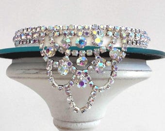 May include: A sparkling, ornate collar necklace with iridescent rhinestones. The necklace features a layered design with a central, dangling pendant. The rhinestones are set in a silver-tone metal, creating a glamorous and eye-catching accessory.