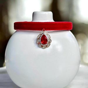 May include: A red velvet choker necklace with a teardrop-shaped pendant featuring a large red gemstone surrounded by clear rhinestones.