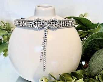 May include: A gray pet collar with a sparkling rhinestone design. The collar features a bow-shaped centerpiece and a dangling strand of rhinestones. The collar is displayed on a white vase with green foliage in the background.