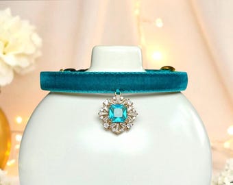 Puede incluir: Un collar de terciopelo turquesa con un colgante brillante. El colgante presenta una gema cuadrada de color azul claro rodeada de cristales transparentes más pequeños. El collar se muestra sobre una superficie blanca y redondeada.
