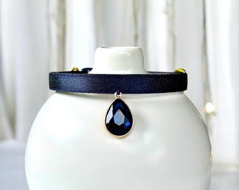 May include: A navy blue choker necklace with a teardrop-shaped, dark blue pendant. The pendant is set in a gold-colored metal. The choker is displayed on a white ceramic object against a soft, blurred background.