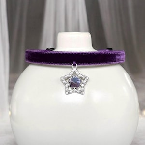 May include: A purple velvet choker necklace with a silver star pendant. The star pendant is encrusted with clear crystals and a central purple gemstone. The choker is displayed on a white, rounded surface.